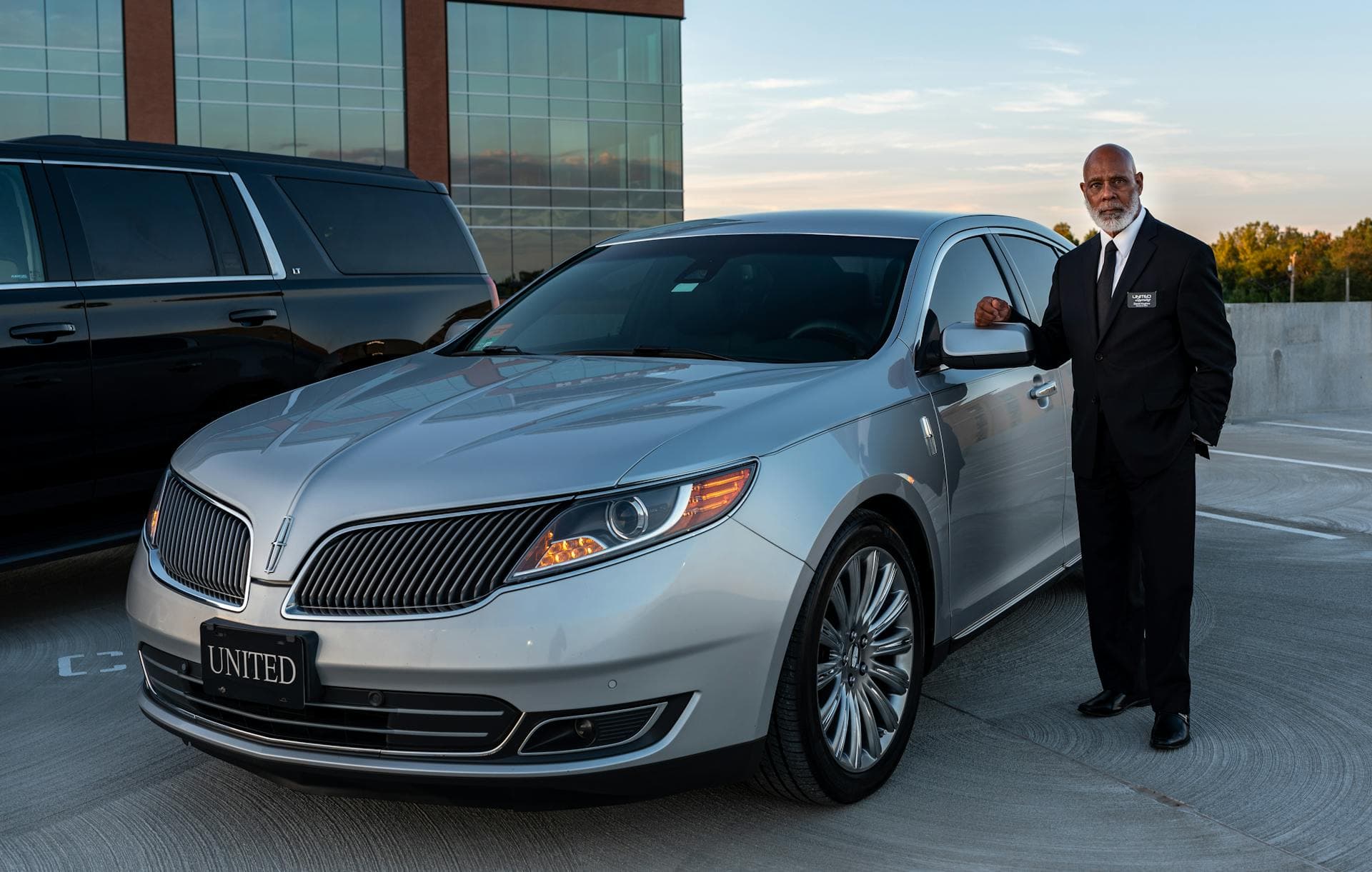 Chauffeur in suit standing beside luxury chauffeured vehicle, owner-operated service detail