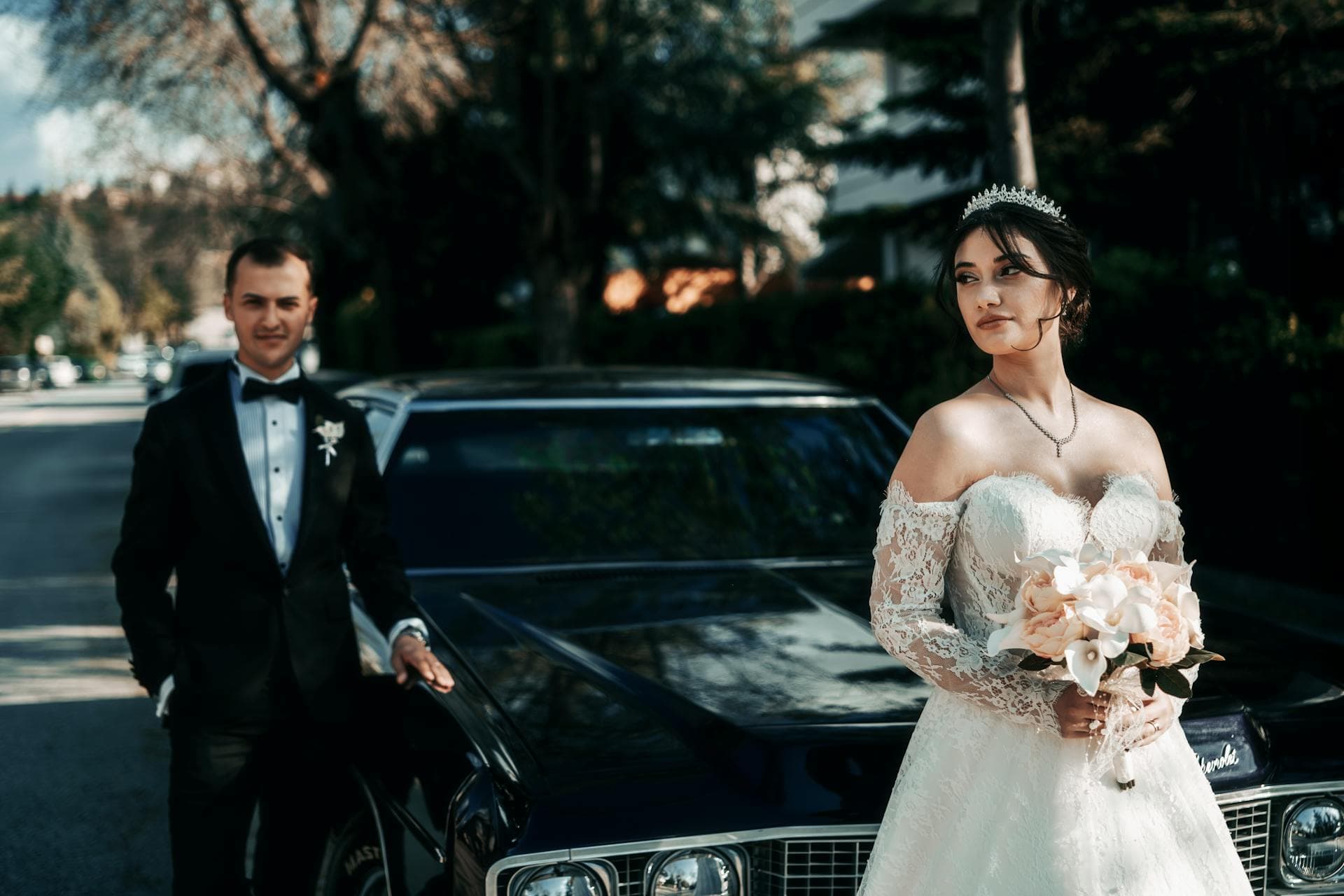 Bride in lace gown with bouquet posing alongside groom beside classic wedding car, Adelaide bridal service